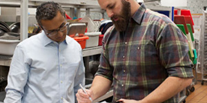 Two people in a kitchen setting near metal shelves with utensils. Two people in a kitchen setting near metal shelves with utensils.