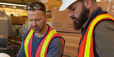 Two people in safety vests are seen in a warehouse setting with stacks of wooden planks behind them. Two people in safety vests are seen in a warehouse setting with stacks of wooden planks behind them.