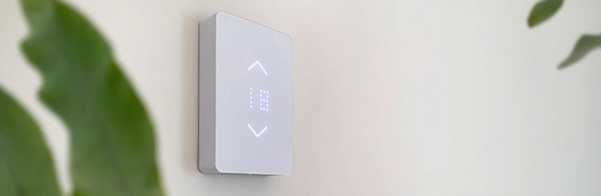 Mysa thermostat on the wall and plant. Mysa smart thermostat on a white wall in the background with part of a plant leaf in the foreground.