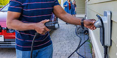 A man plugs an electric charger. The background shows his interracial family walking around and approaching.