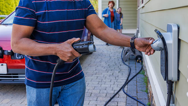 A man plugs an electric charger. The background shows his interracial family walking around and approaching.