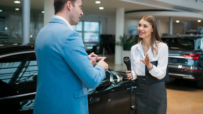 Sales manager holding car charging adapter and explaining the features of an electric car in showroom.