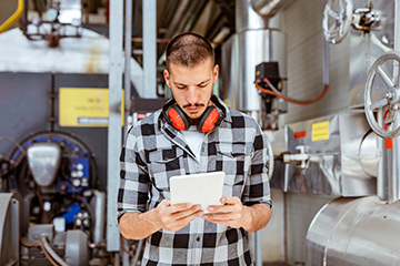 Maintenance engineer stands in the basement of a power station, inspecting the heating system on a digital tablet